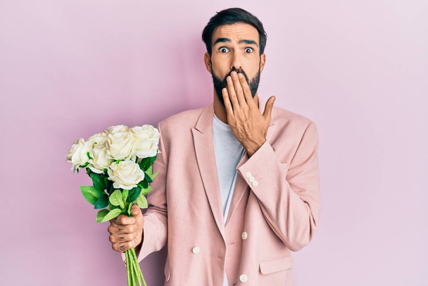 Junge hispanische Mann mit Blumen bedeckt Mund mit der Hand, schockiert und Angst vor Fehler. Überraschte Miene  - Foto, Bild