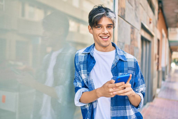 Молодой латиноамериканец улыбается счастливо, используя смартфон в городе. - Фото, изображение