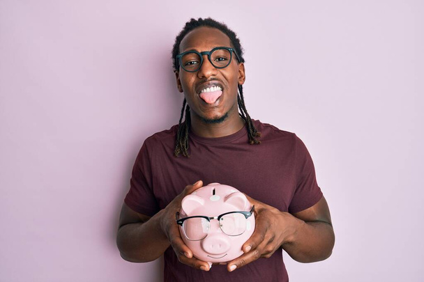 African american man with braids holding piggy bank with glasses sticking tongue out happy with funny expression.  - Fotó, kép