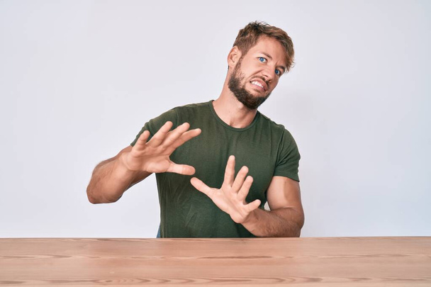 Junge kaukasische Mann in lässiger Kleidung auf dem Tisch sitzen angewiderte Miene, ungehalten und ängstlich dabei Ekel Gesicht, weil Abneigung Reaktion. mit erhobenen Händen  - Foto, Bild