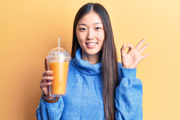 Jeune belle femme chinoise boire un verre de jus d'orange faire ok signe avec les doigts, sourire geste amical excellent symbole  - Photo, image