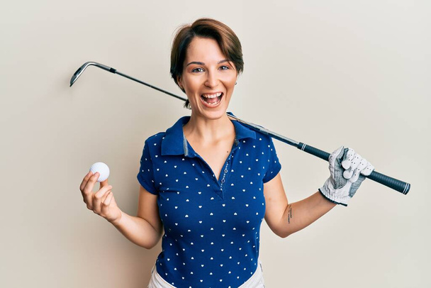 Giovane donna bruna con i capelli corti che tiene palla e golf club sorridente e ridente forte perché divertente scherzo pazzo.  - Foto, immagini