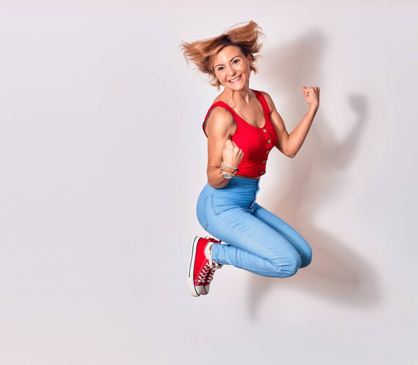 Young beautiful woman wearing casual clothes smiling happy. Jumping with smile on face doing winner gesture with thumbs up over isolated white background - Zdjęcie, obraz