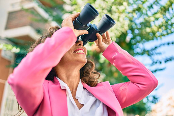 Middle age hispanic businesswoman smiling happy looking for new opportunity using binoculars at the city. - Photo, image