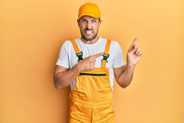 Young handsome man wearing handyman uniform over yellow background pointing aside worried and nervous with both hands, concerned and surprised expression  - Photo, Image