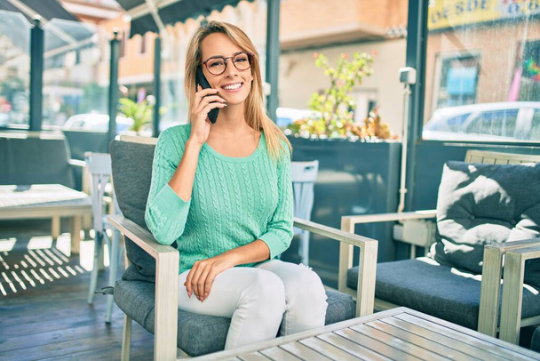 jong blond vrouw glimlachen gelukkig praten op de smartphone zitten op de coffeeshop terras. - Foto, afbeelding