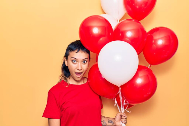 Mladá žena drží balónky vyděšené a ohromený s otevřenými ústy pro překvapení, nevěřící tvář  - Fotografie, Obrázek