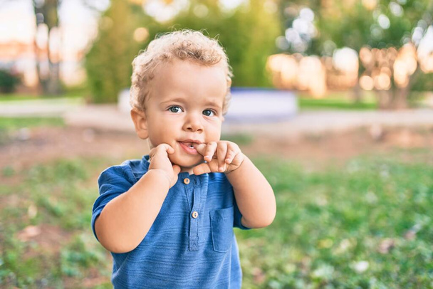 Smutny chłopczyk dotykający dziąseł palcami, bo w słoneczny dzień w parku boli go ząb. piękne blond włosy mężczyzna maluch w ból dla nowych zębów dziecka na zewnątrz - Zdjęcie, obraz