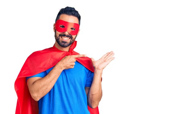Mladý hispánec muž na sobě super hrdina kostým ohromen a usmívá se na kameru při prezentaci s rukou a ukazuje prstem.  - Fotografie, Obrázek