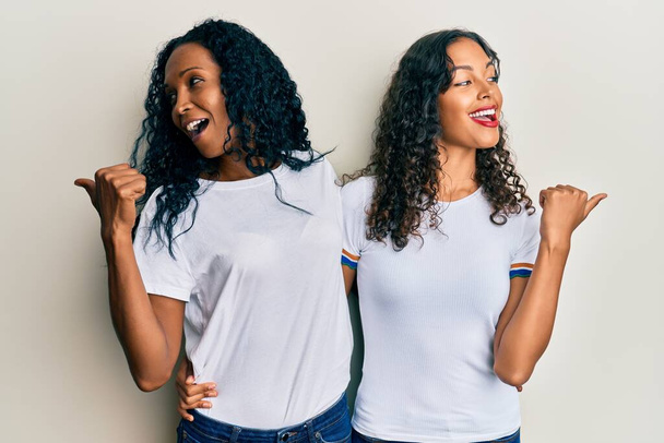 Afroamerikanische Mutter und Tochter tragen lässiges weißes T-Shirt, das den Daumen zur Seite zeigt und glücklich mit offenem Mund lächelt  - Foto, Bild