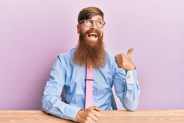 Молодой ирландский рыжий мужчина в деловой рубашке и галстуке сидит на столе улыбаясь со счастливым лицом глядя и указывая в сторону с большим пальцем вверх.  - Фото, изображение