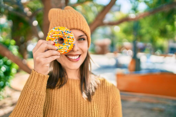 jonge blonde vrouw glimlachen gelukkig holding smakelijke kleurrijke donut op oog in het park. - Foto, afbeelding