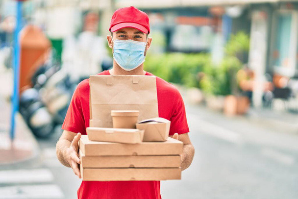 Joven repartidor caucásico con máscara médica sosteniendo comida a domicilio en la ciudad. - Foto, imagen