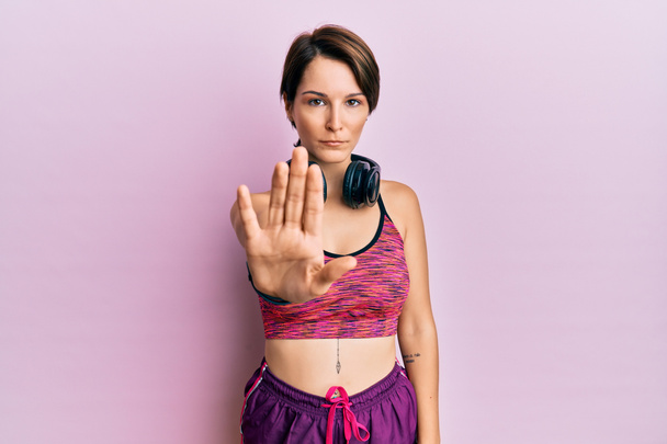 Young brunette woman with short hair wearing sportswear and headphones doing stop sing with palm of the hand. warning expression with negative and serious gesture on the face.  - Foto, Bild