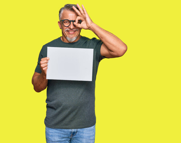 Grauhaariger Mann mittleren Alters, der ein leeres Banner hält und glücklich lächelt, während er ein Zeichen macht, auf dem die Hand auf dem Auge durch Finger schaut  - Foto, Bild