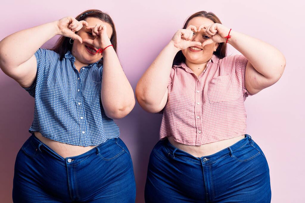 Young plus size twins wearing casual clothes doing heart shape with hand and fingers smiling looking through sign  - Foto, imagen
