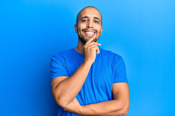 Ein hispanischer erwachsener Mann in lässigem blauem T-Shirt lächelt selbstbewusst in die Kamera, die Arme verschränkt und die Hand am Kinn. Positives Denken.  - Foto, Bild