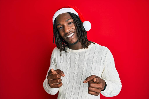 Африканский американец с косичками в рождественской шляпе указывает пальцем на камеру со счастливым и смешным лицом. хорошая энергия и вибрации.  - Фото, изображение