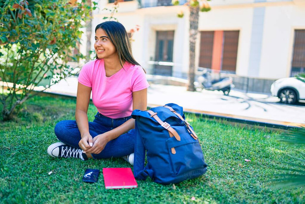 Genç Latin kız gülümsüyor. Üniversite kampüsünde çimlerin üzerinde mutlu mesut oturuyor. - Fotoğraf, Görsel