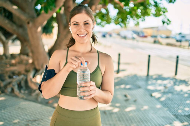 Jonge blonde sportvrouw doet oefening drinken fles water in de stad. - Foto, afbeelding