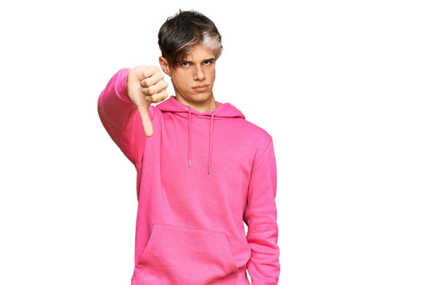 Молодой латиноамериканец в обычной розовой толстовке, выглядящий несчастным и злым, демонстрирующий отказ и негатив с опущенными пальцами. плохое выражение.  - Фото, изображение