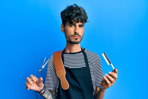 Молодий іспаномовний чоловік у бар'єрному фартусі тримає бритву та ножиці, розслаблений серйозним виразом обличчя. простий і природний погляд на камеру
.  - Фото, зображення