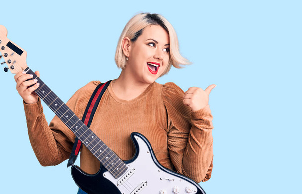 Junge blonde Plus-Size-Frau spielt E-Gitarre und zeigt Daumen zur Seite, lächelt glücklich mit geöffnetem Mund  - Foto, Bild