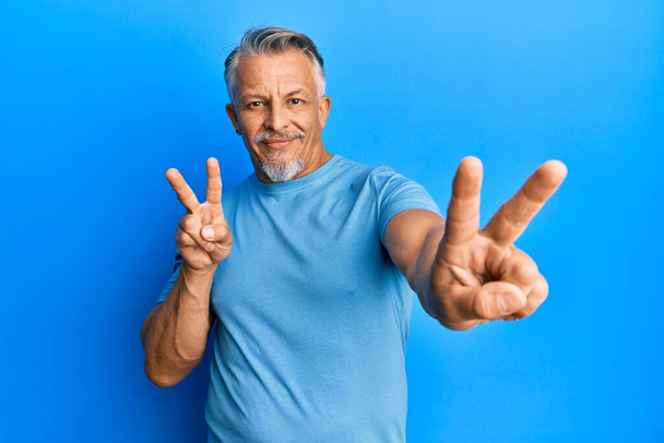 Μεσήλικας γκριζομάλλης άντρας που φοράει καθημερινά ρούχα χαμογελώντας κοιτάζοντας στην κάμερα που δείχνει δάχτυλα να κάνουν το σήμα της νίκης. Νούμερο δύο..  - Φωτογραφία, εικόνα