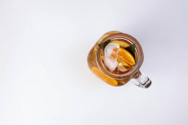 Liquid ice lemon orange tea with slice green leaf cinnamon stick in transparent glass jar mug on white background top view - Foto, Bild
