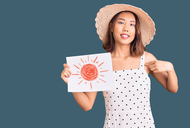 Yazlık şapka takan genç ve güzel bir Çinli kız elinde güneş olan mutlu ve gururlu bir kişiyi işaret ediyor.  - Fotoğraf, Görsel