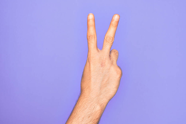 Hand of caucasian young man showing fingers over isolated purple background counting number 2 showing two fingers, gesturing victory and winner symbol - Photo, Image