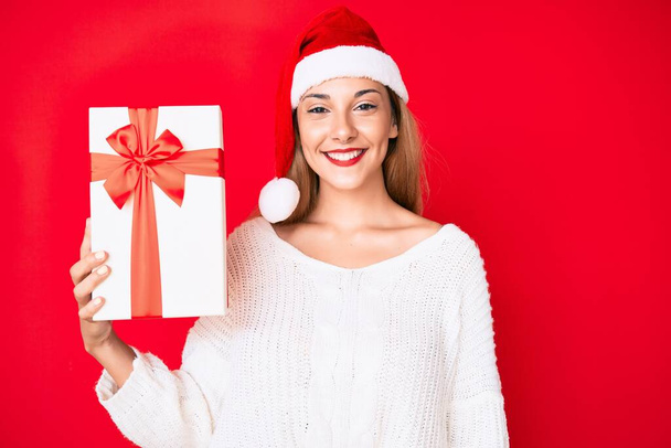Genç esmer kadın Noel şapkası takıyor ve elinde bir hediye tutuyor. Pozitif ve mutlu görünüyor. Kendine güvenen bir gülümsemeyle dişlerini gösteriyor.  - Fotoğraf, Görsel
