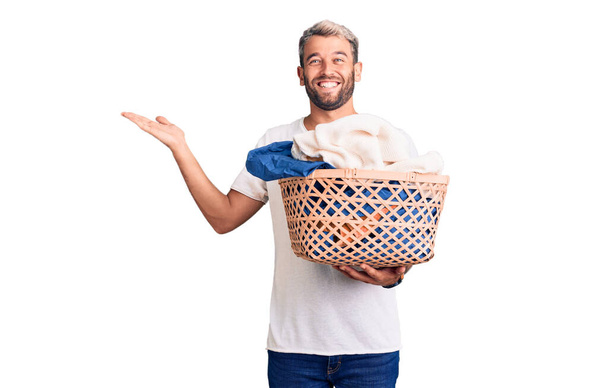 Joven hombre rubio guapo sosteniendo cesta de ropa con ropa celebrando la victoria con sonrisa feliz y la expresión ganadora con las manos levantadas  - Foto, Imagen