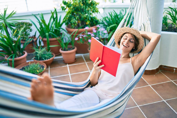 jong mooi blank vrouw glimlachen gelukkig rusten op een hangmat het lezen van een boek thuis - Foto, afbeelding