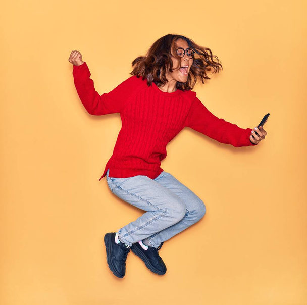 Young beautiful latin woman wearing glasses smiling happy. Jumping with smile on face make selfie by smartphone over isolated yellow background - Photo, Image