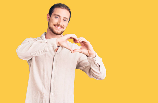 Jeune homme caucasien beau portant des vêtements décontractés souriant dans l'amour faisant forme de symbole de coeur avec les mains. concept romantique.  - Photo, image
