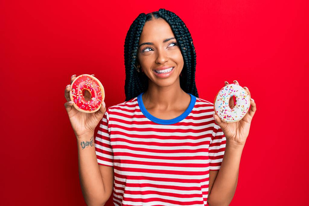 Schöne hispanische Frau mit leckeren bunten Donuts lächelnd zur Seite schauen und wegstarren denken.  - Foto, Bild