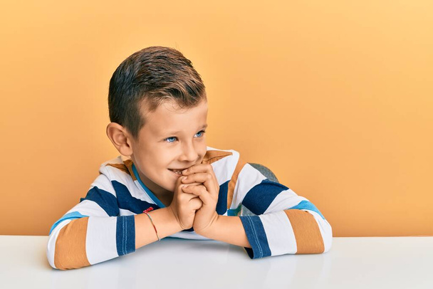 Entzückendes kaukasisches Kind in lässiger Kleidung, das nervös und aufgeregt auf dem Tisch sitzt, die Hände am Kinn zur Seite gerichtet  - Foto, Bild