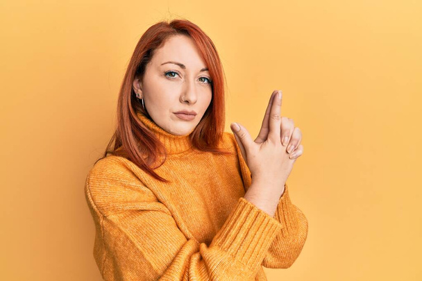Beautiful redhead woman wearing casual winter sweater over yellow background holding symbolic gun with hand gesture, playing killing shooting weapons, angry face  - Photo, Image