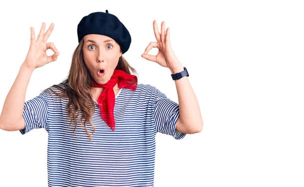 Giovane bella donna bionda con berretto francese e t-shirt a righe cercando sorpreso e scioccato facendo ok simbolo di approvazione con le dita. espressione folle  - Foto, immagini