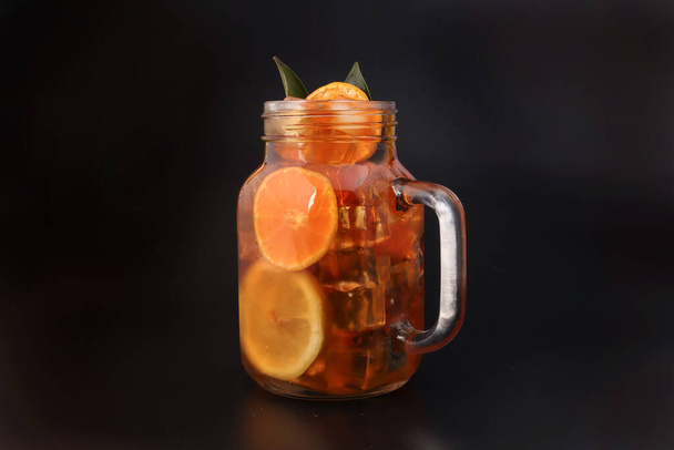 Liquid ice lemon orange tea with slice green leaf cinnamon stick in transparent glass jar mug on black background - Photo, Image