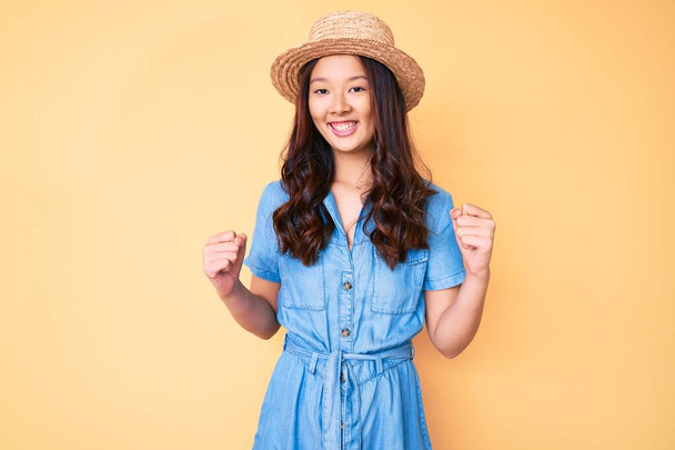 Young beautiful chinese girl wearing summer hat celebrating surprised and amazed for success with arms raised and open eyes. winner concept.  - Фото, изображение