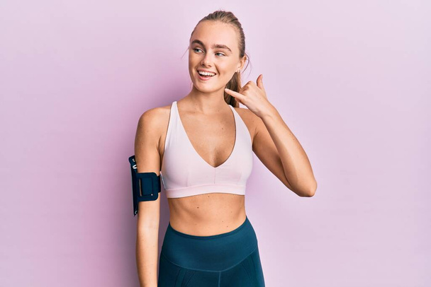 Beautiful blonde woman wearing sportswear and arm band smiling doing phone gesture with hand and fingers like talking on the telephone. communicating concepts.  - Photo, Image