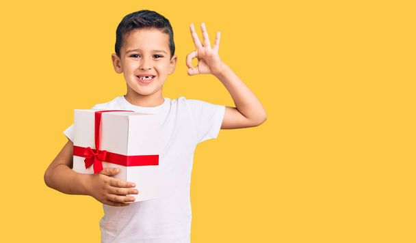 Pequeño niño lindo sosteniendo regalo haciendo signo ok con los dedos, sonriendo gesto amistoso excelente símbolo  - Foto, imagen
