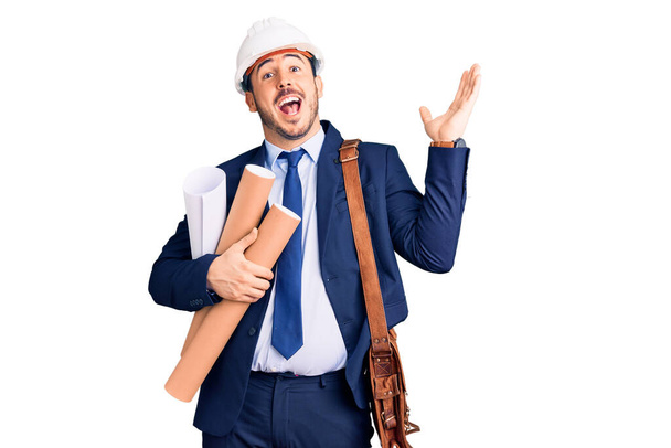 Jonge Spaanse man met een architectenhelm en leren tas met blauwdrukken die de overwinning vieren met een vrolijke glimlach en een winnaarsuitdrukking met opgeheven handen  - Foto, afbeelding