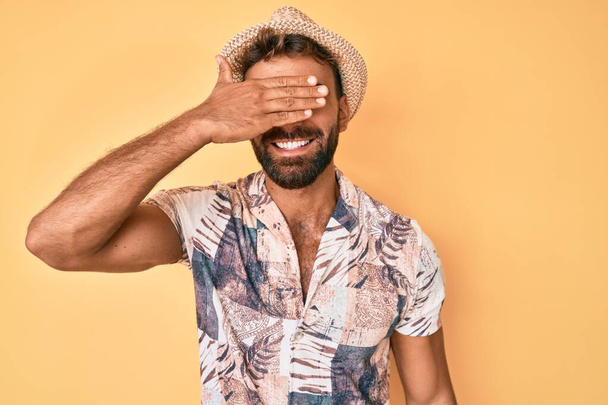 Junge hispanische Mann mit Sommermütze lächelnd und lachend mit der Hand auf dem Gesicht bedeckt Augen für Überraschung. Blindes Konzept.  - Foto, Bild
