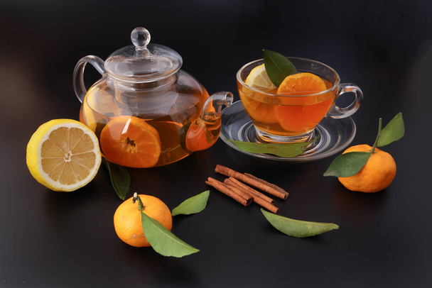 Liquid tea lemon orange slice green leaf cinnamon stick in transparent glass teacup saucer teapot kettle on black background - Photo, image