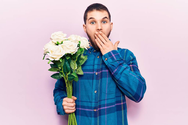 Giovane bell'uomo che tiene mazzo di fiori che coprono la bocca con mano, scioccato e spaventato per errore. espressione sorpresa  - Foto, immagini
