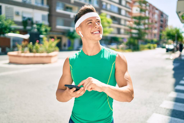 Young hispanic sportsman using smartphone and earphones walking at street of city - Photo, Image