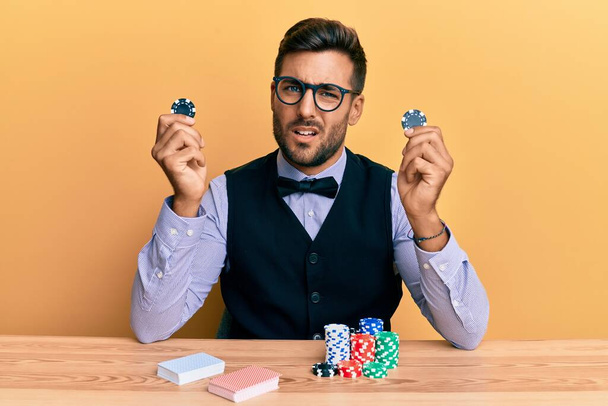 Handsome hispanic croupier man sitting on the table holding casino chips clueless and confused expression. doubt concept.  - Photo, Image